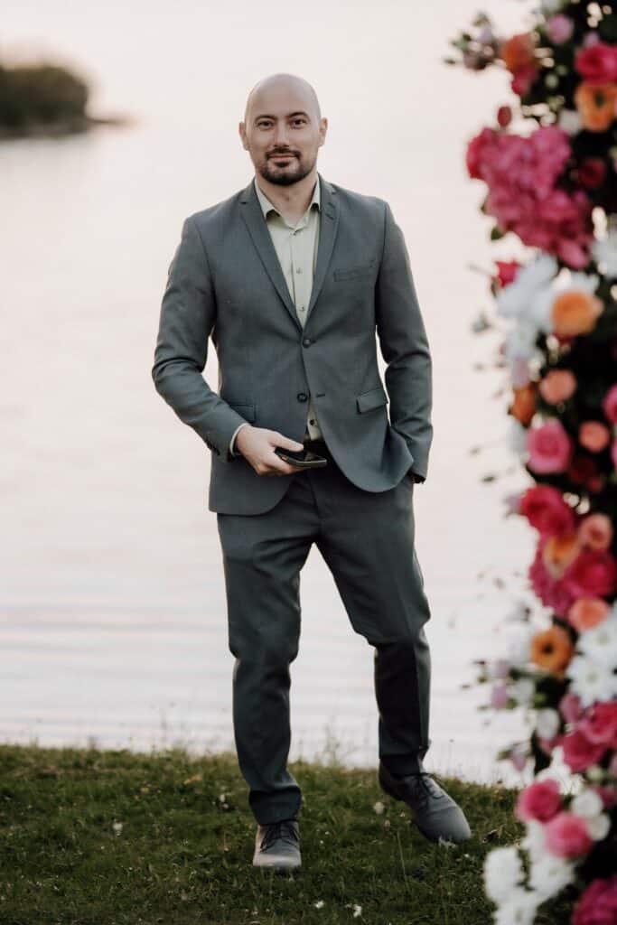 DJ im grauen Anzug auf Wiese vor einem See mit Handy in der Hand.